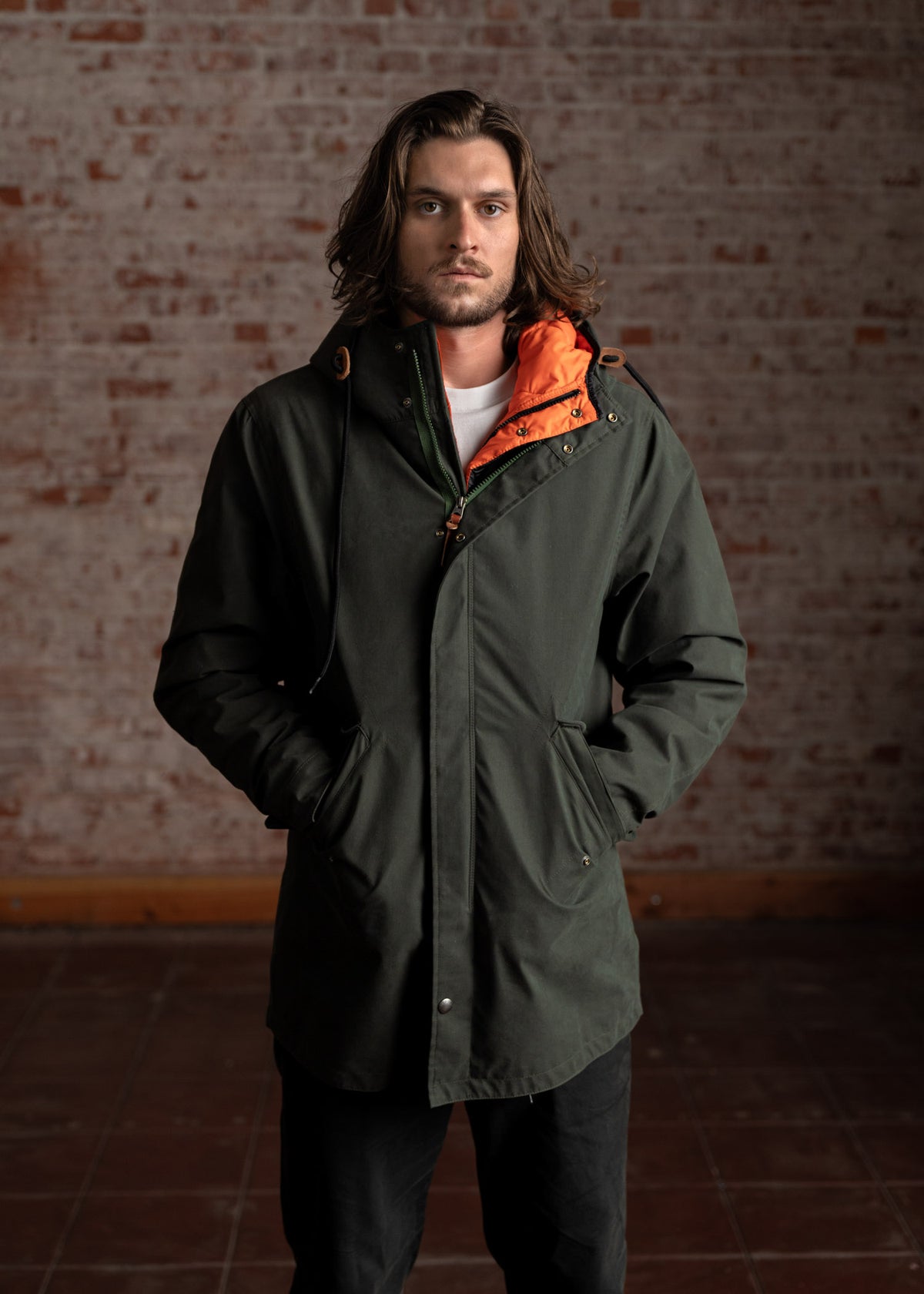 Man in a green parka with orange lining stands against a rustic brick wall, hands in pockets, exuding a casual, rugged style.
