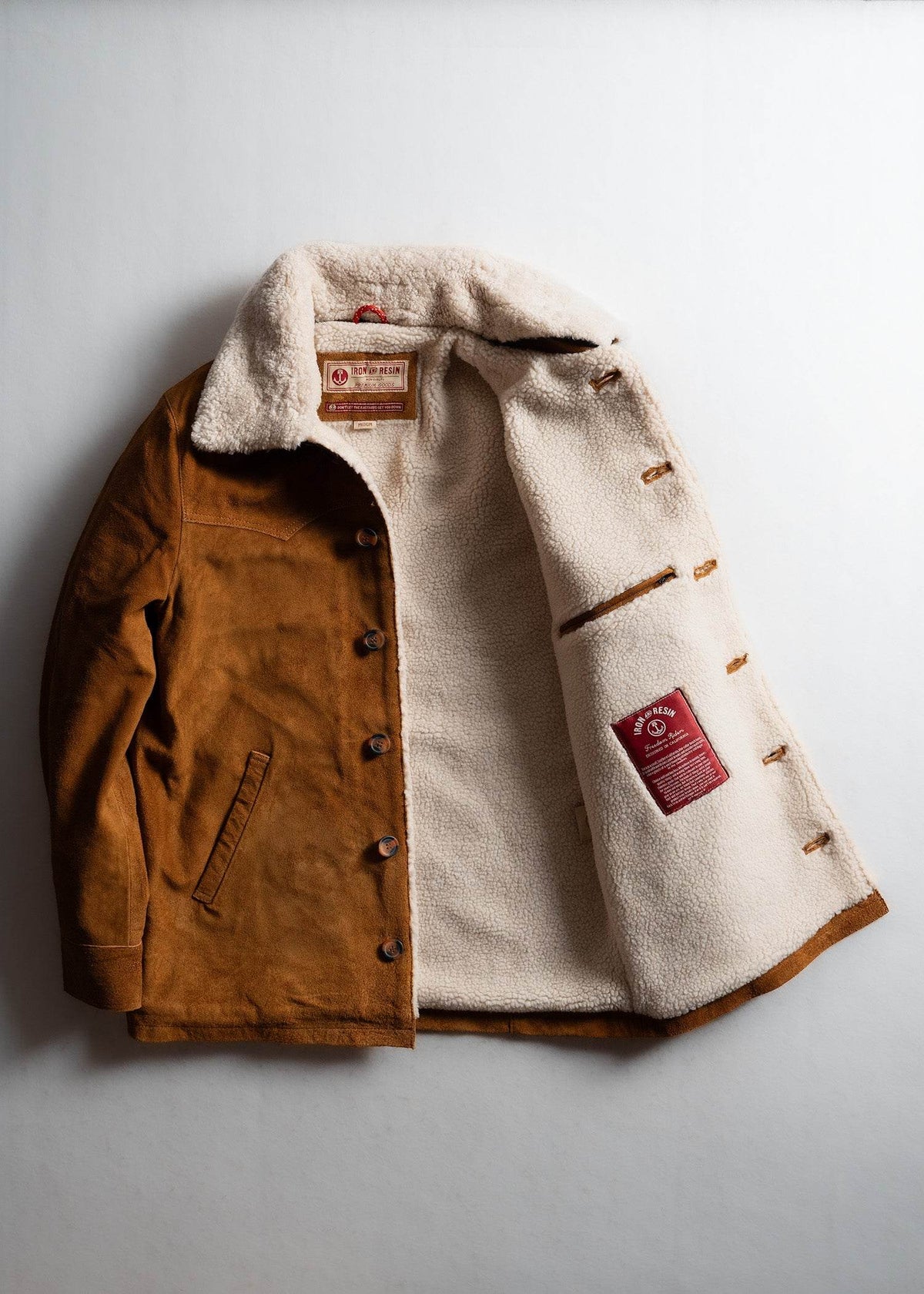 Brown suede jacket with white fleece lining, open to show interior label. Cozy and stylish outerwear on a plain background.