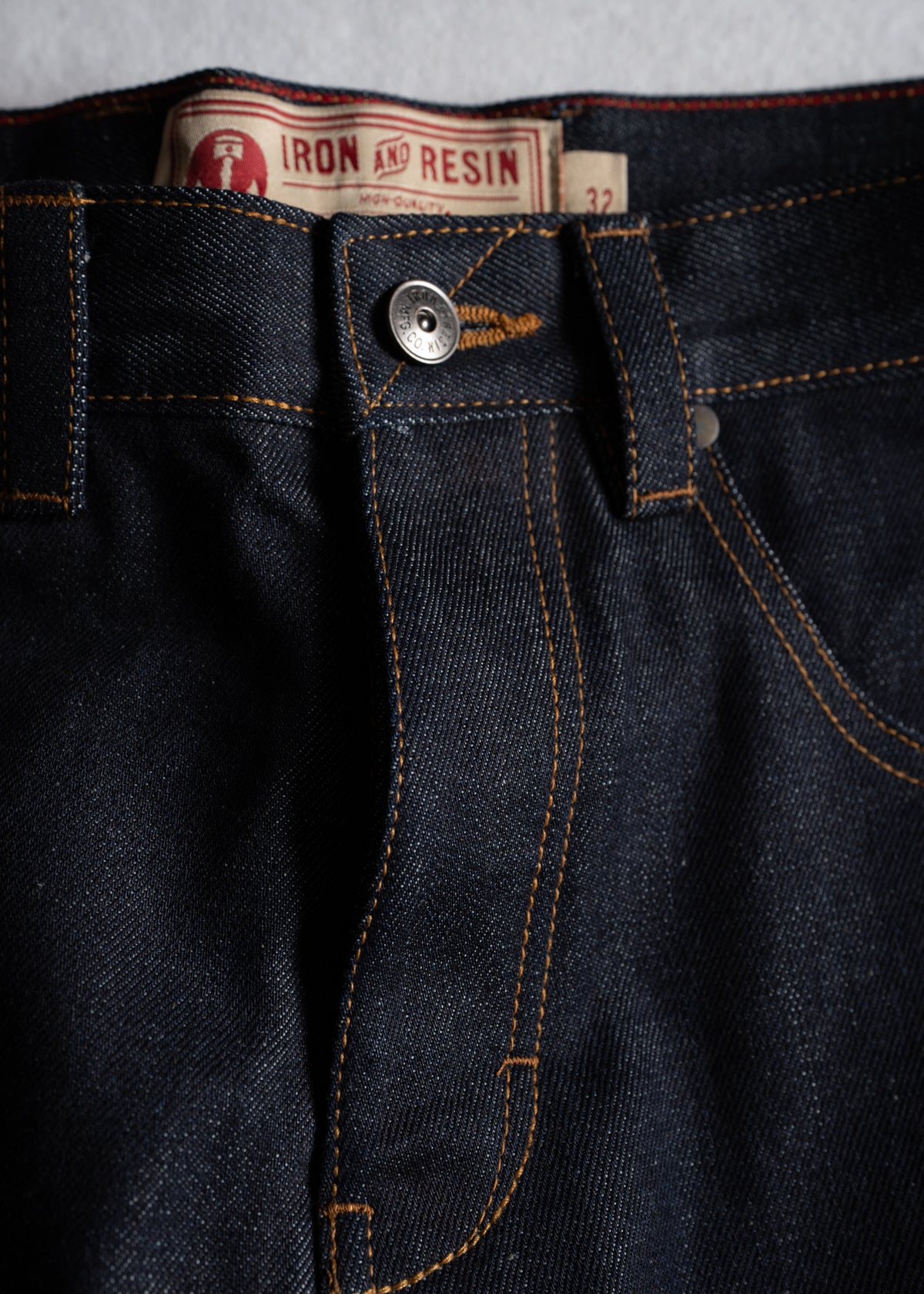 Close-up of dark denim jeans with orange stitching and a metal button. The label reads "Iron and Resin" with size 32.