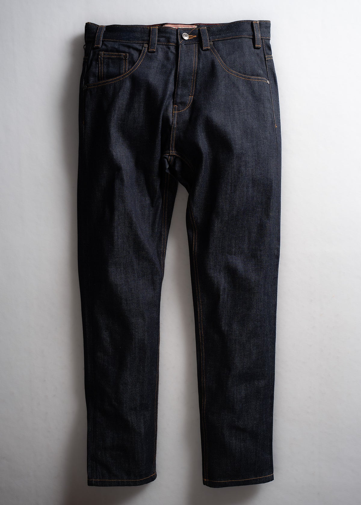 Dark blue jeans with orange stitching hang against a light background, showcasing classic pockets and button closure.