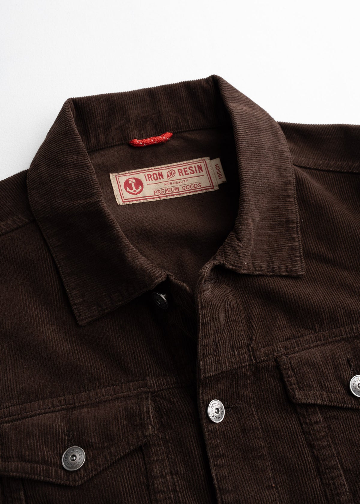 Close-up of a brown corduroy jacket with Iron and Resin label, featuring metal buttons and a classic collar on a white background.
