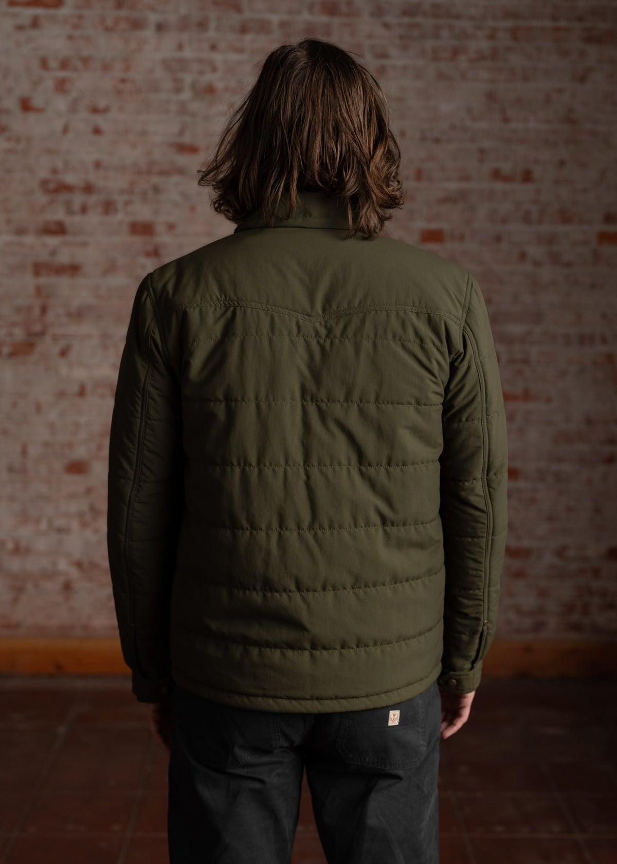 A person with long hair, wearing a green quilted jacket, stands facing a brick wall in a dimly lit room.