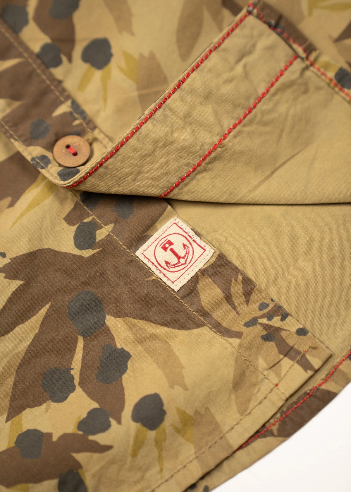Close-up of a camouflage shirt with red stitching and a button, featuring a small red logo tag on the fabric.
