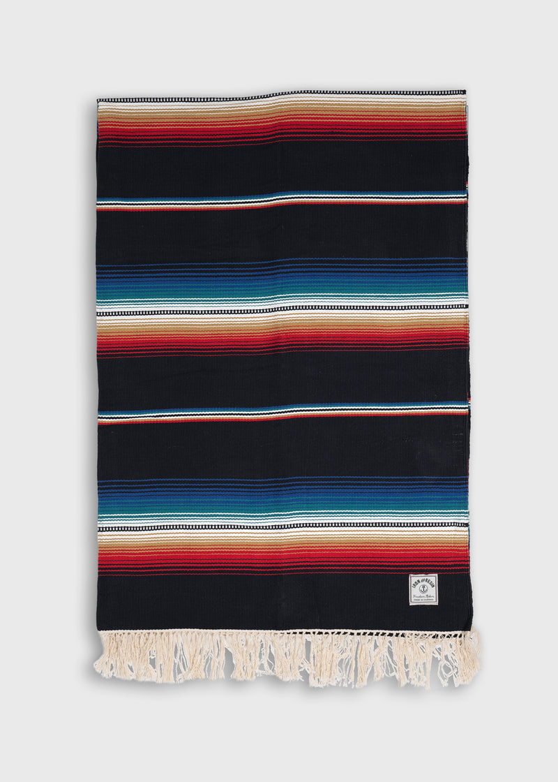Colorful striped blanket with fringe edges, featuring bold red, blue, and black patterns. A small label is visible on one corner.