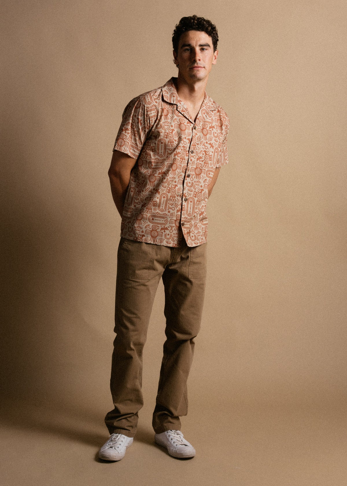 Man in patterned shirt and khaki pants stands against a beige backdrop, hands behind back, wearing white sneakers.