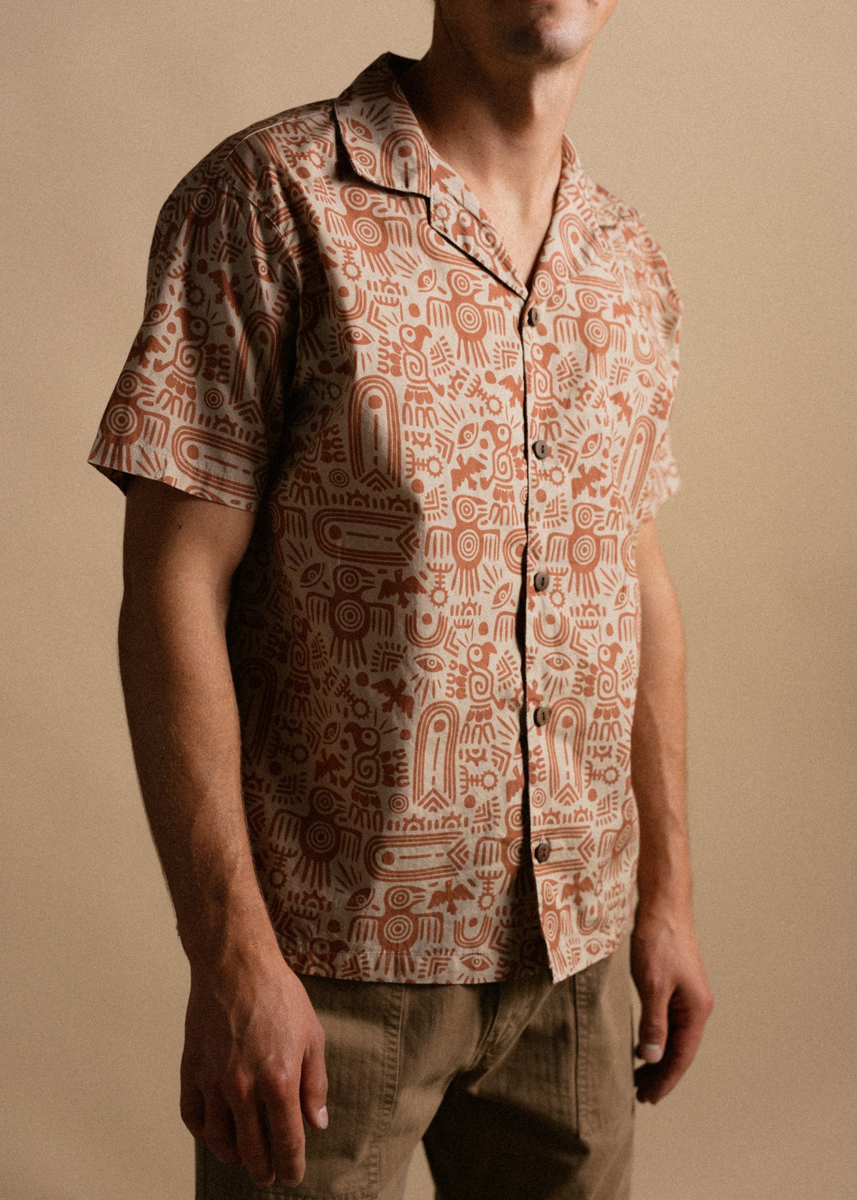 Man wearing a short-sleeve shirt with intricate brown patterns, standing against a beige background.
