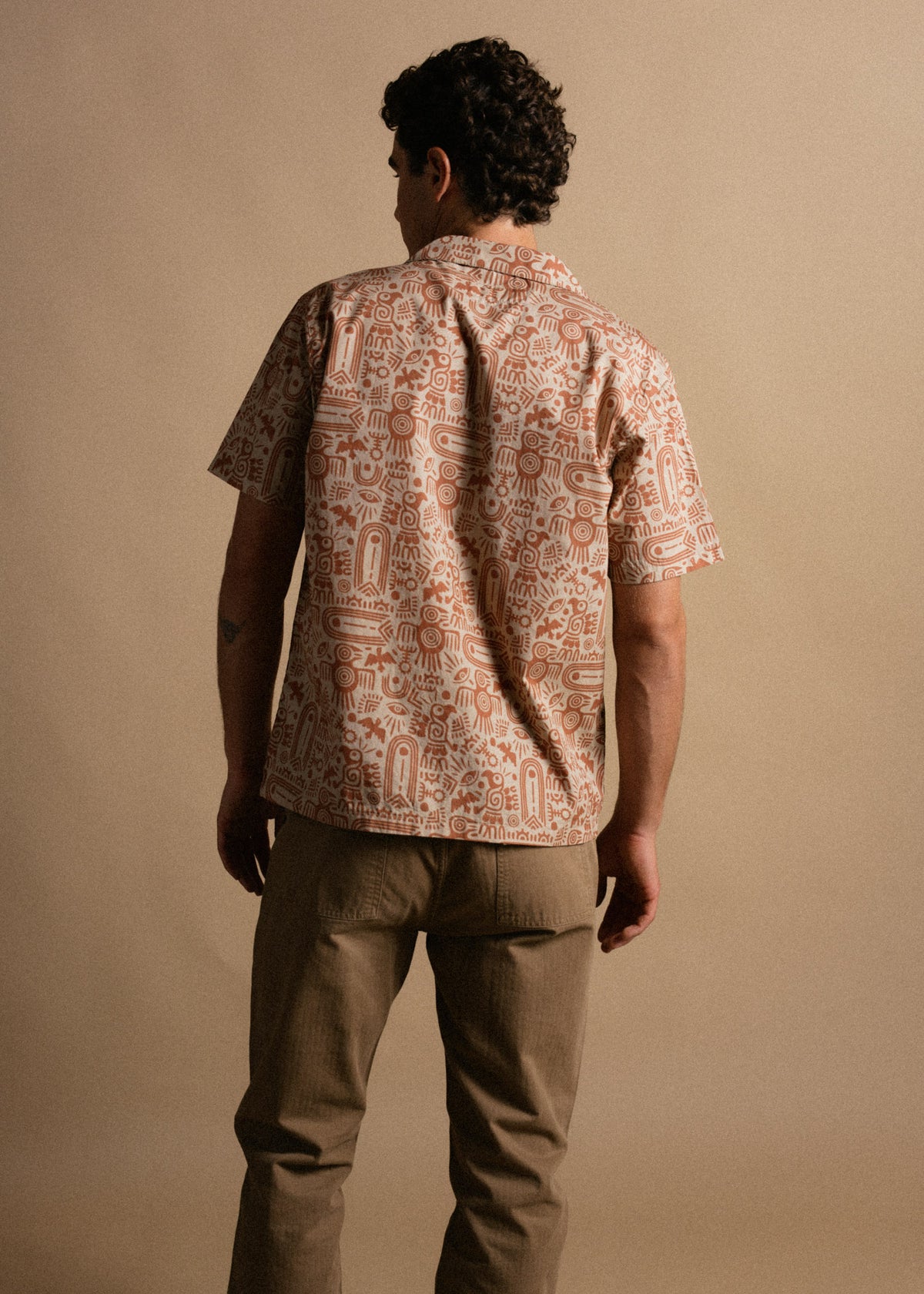 Man in patterned shirt and khaki pants stands against a beige backdrop, facing away. The shirt features intricate, abstract designs.
