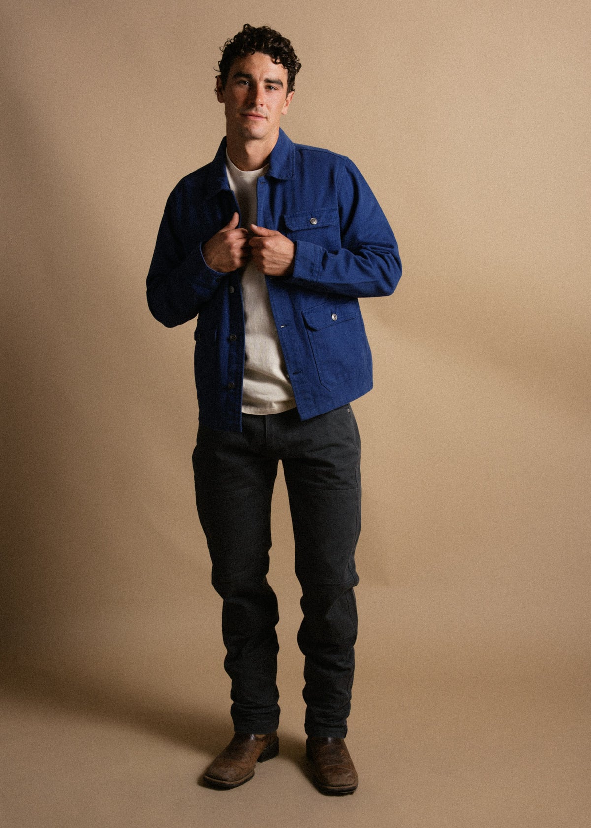 Man in blue jacket, white shirt, dark pants, and brown boots stands against a beige background, hands adjusting jacket.