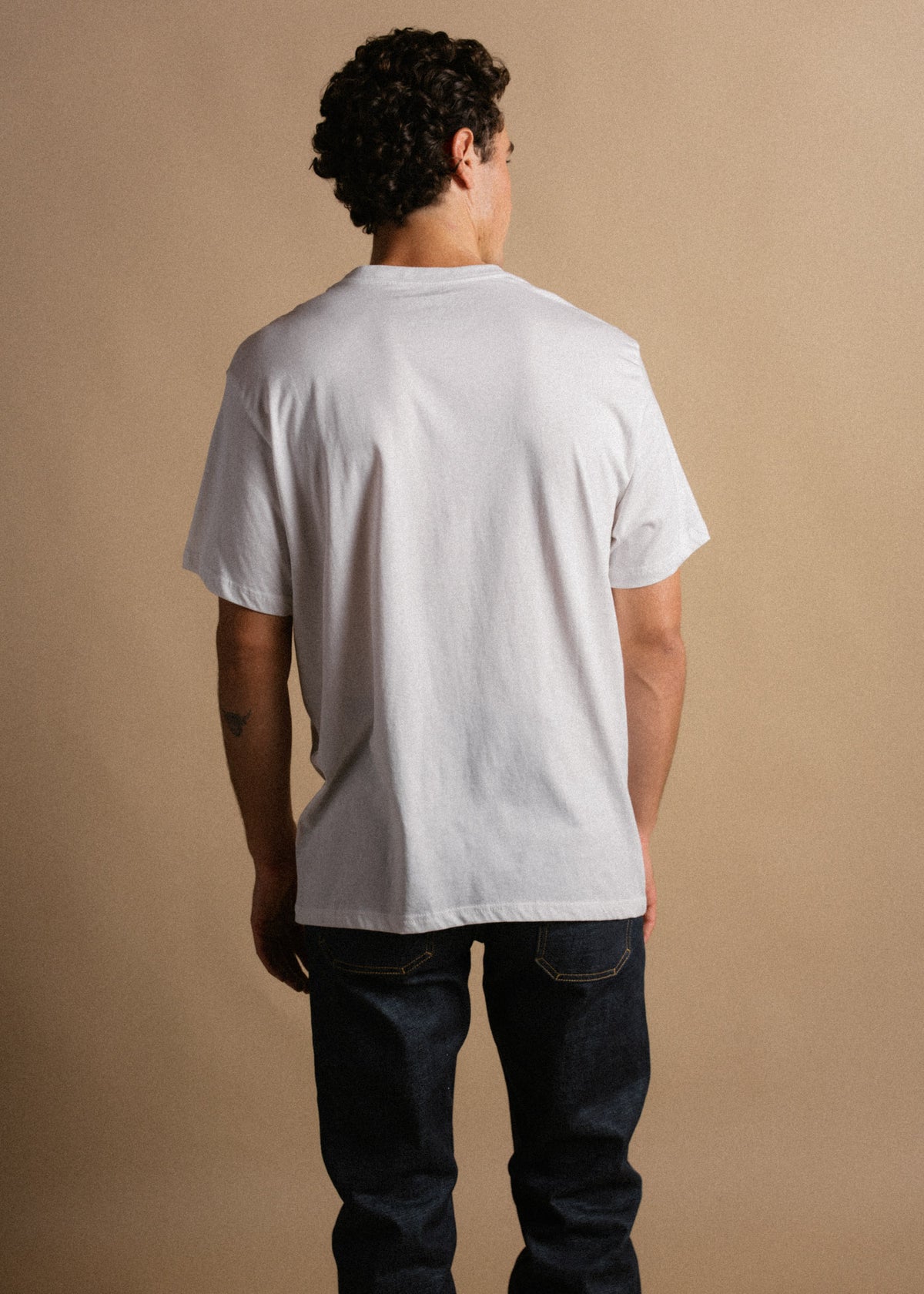 Man in white t-shirt and jeans stands with back to camera against beige background, showcasing casual fashion style.