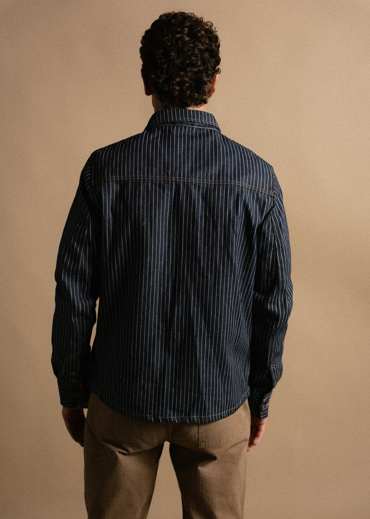 Man in a striped denim shirt and khaki pants stands facing away against a beige background, highlighting casual style.