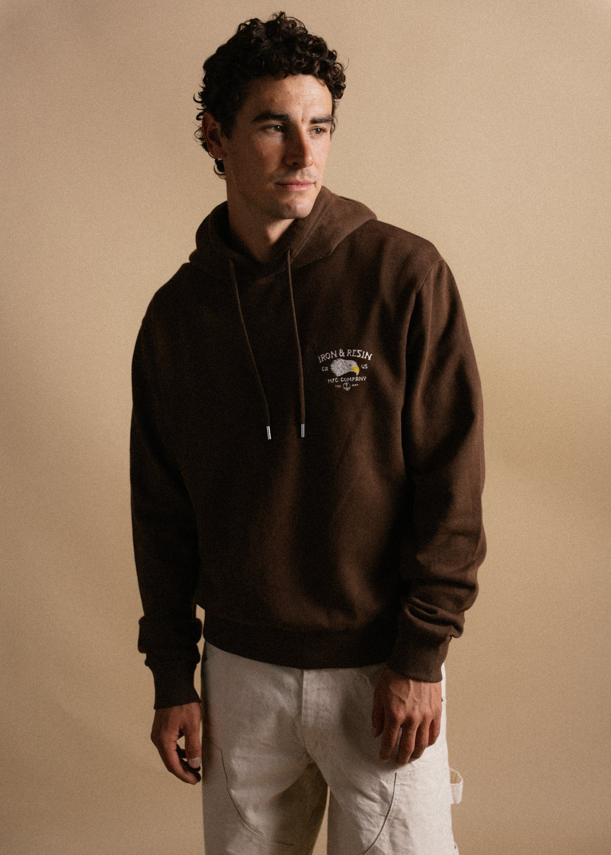 Man in a brown hoodie with "Iron & Resin" logo, stands against a beige background, looking to the side, hands relaxed.