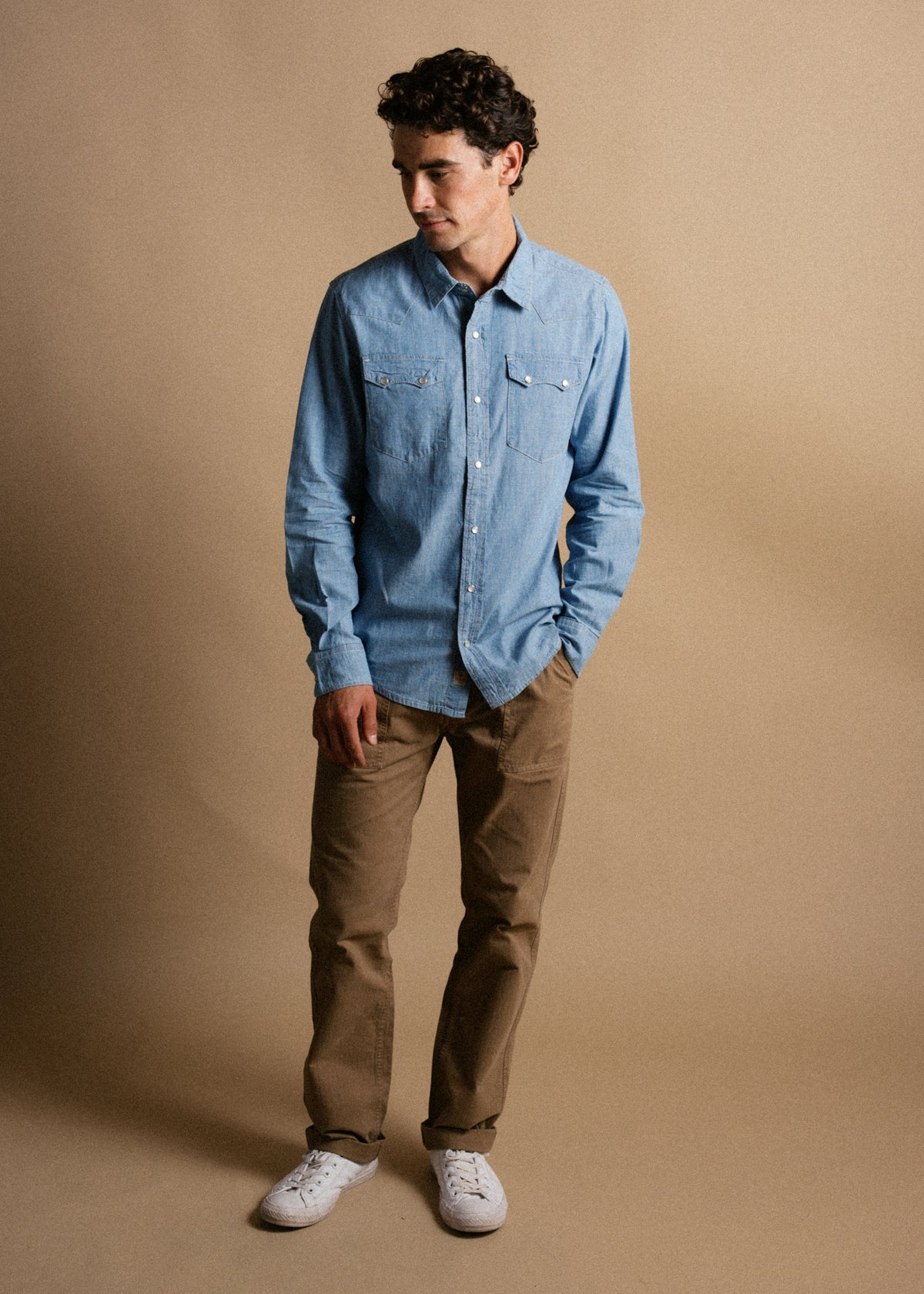 Man in a blue denim shirt and khaki pants stands against a beige background, looking down with a relaxed posture and white sneakers.