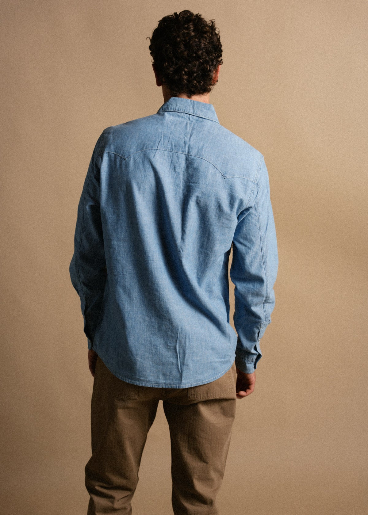 Man in a blue denim shirt and khaki pants stands with his back to the camera against a beige background.