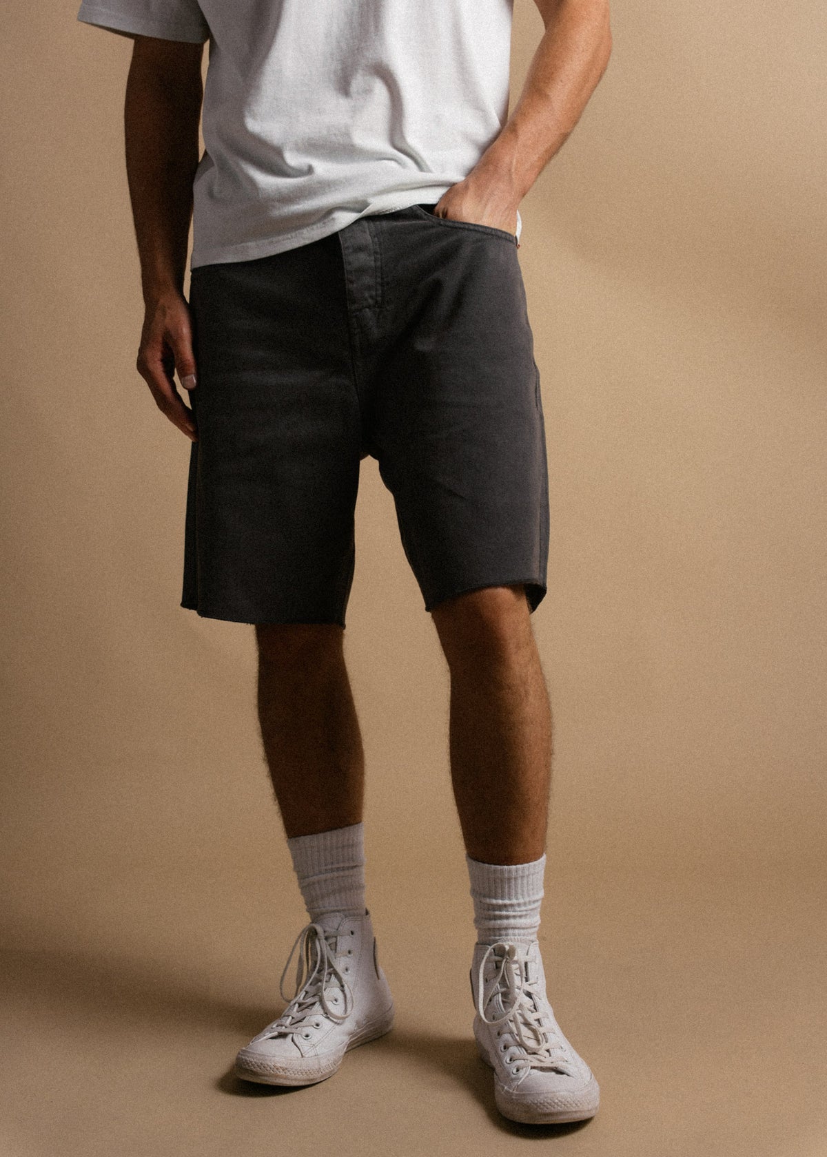 Man in gray shorts and white sneakers stands against a beige background, hands in pockets, casual style.
