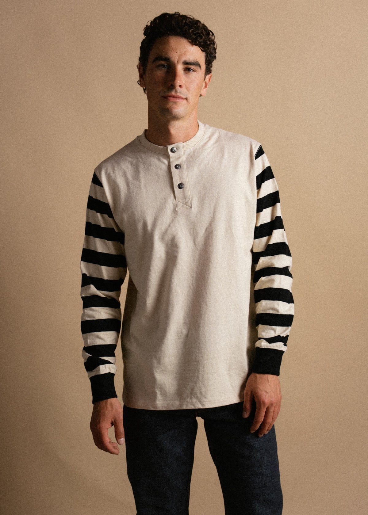 Man in a beige henley with black-striped sleeves stands against a tan background, exuding casual style and confidence.