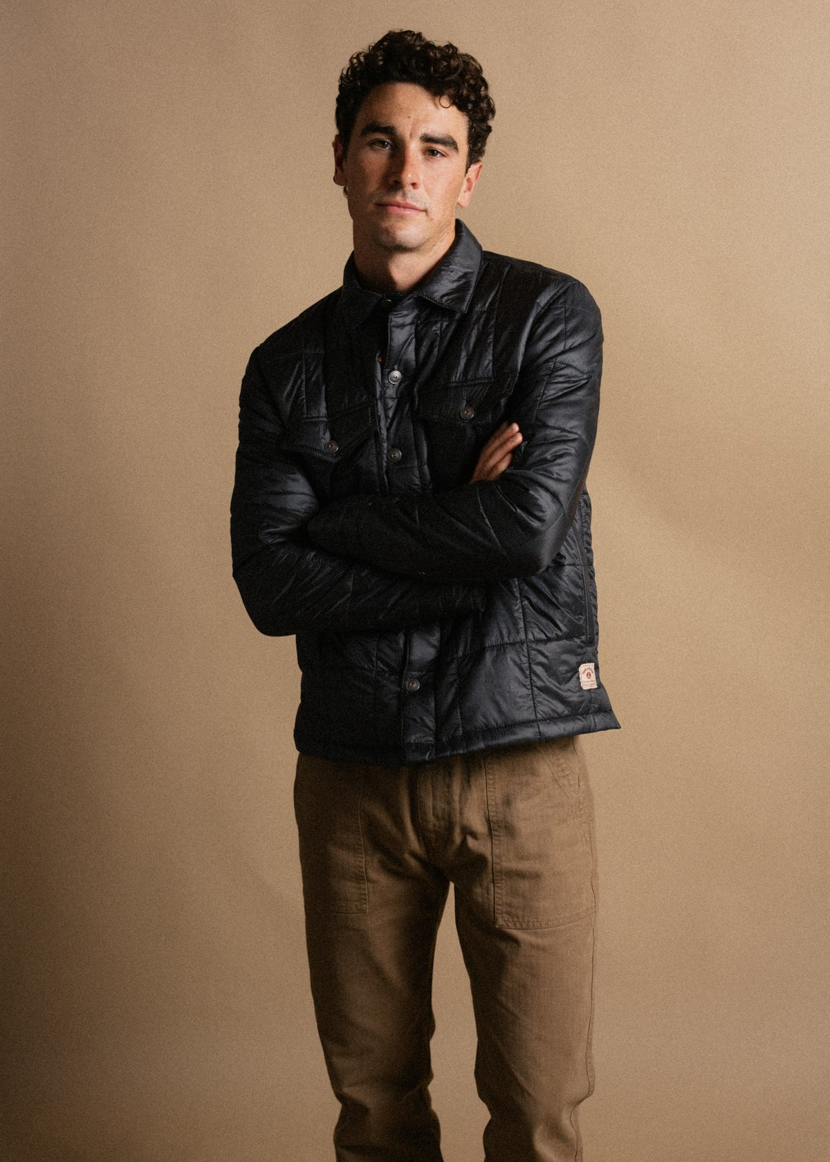 Man in a dark jacket and brown pants stands confidently against a beige background, arms crossed, looking directly at the camera.