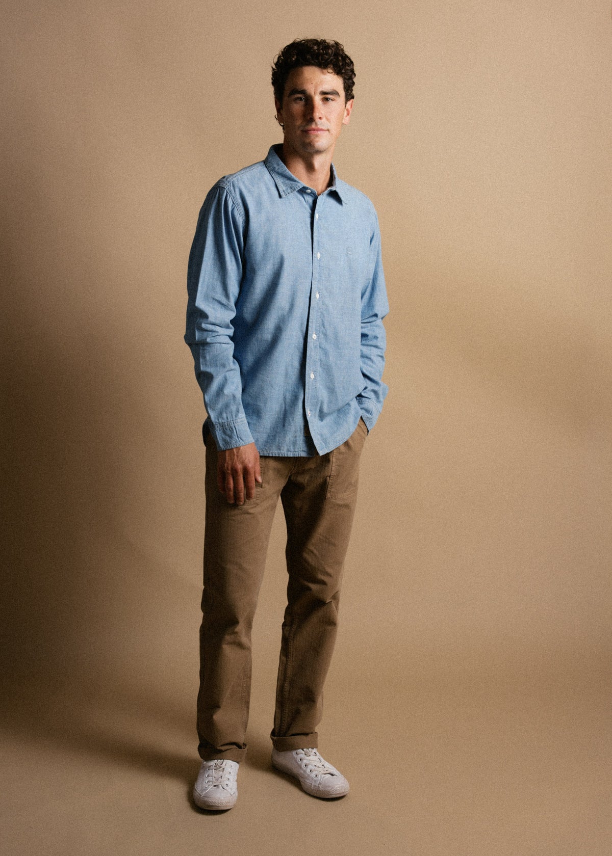 Man in blue shirt and khaki pants stands against a beige background, hands in pockets, wearing white sneakers, looking at camera.