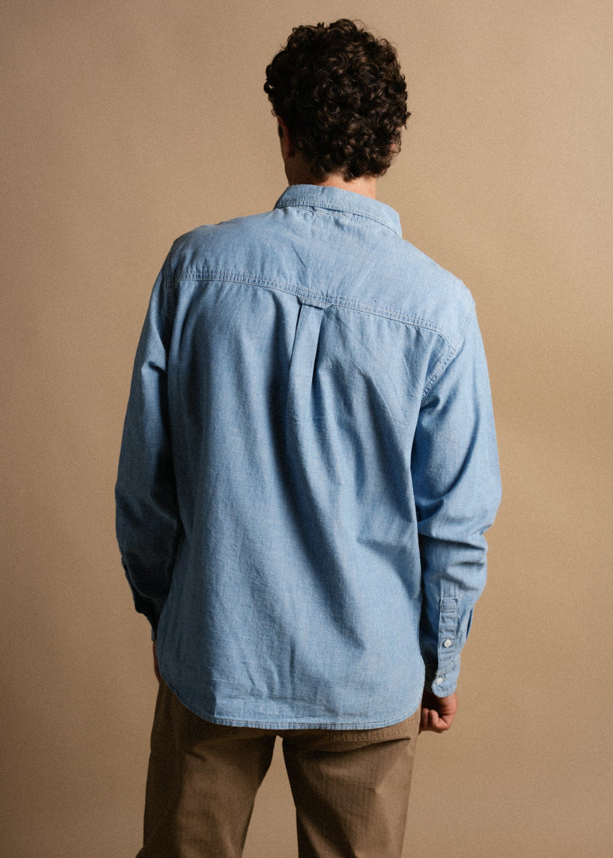 Man in blue denim shirt and brown pants stands with back to camera against a plain beige background.