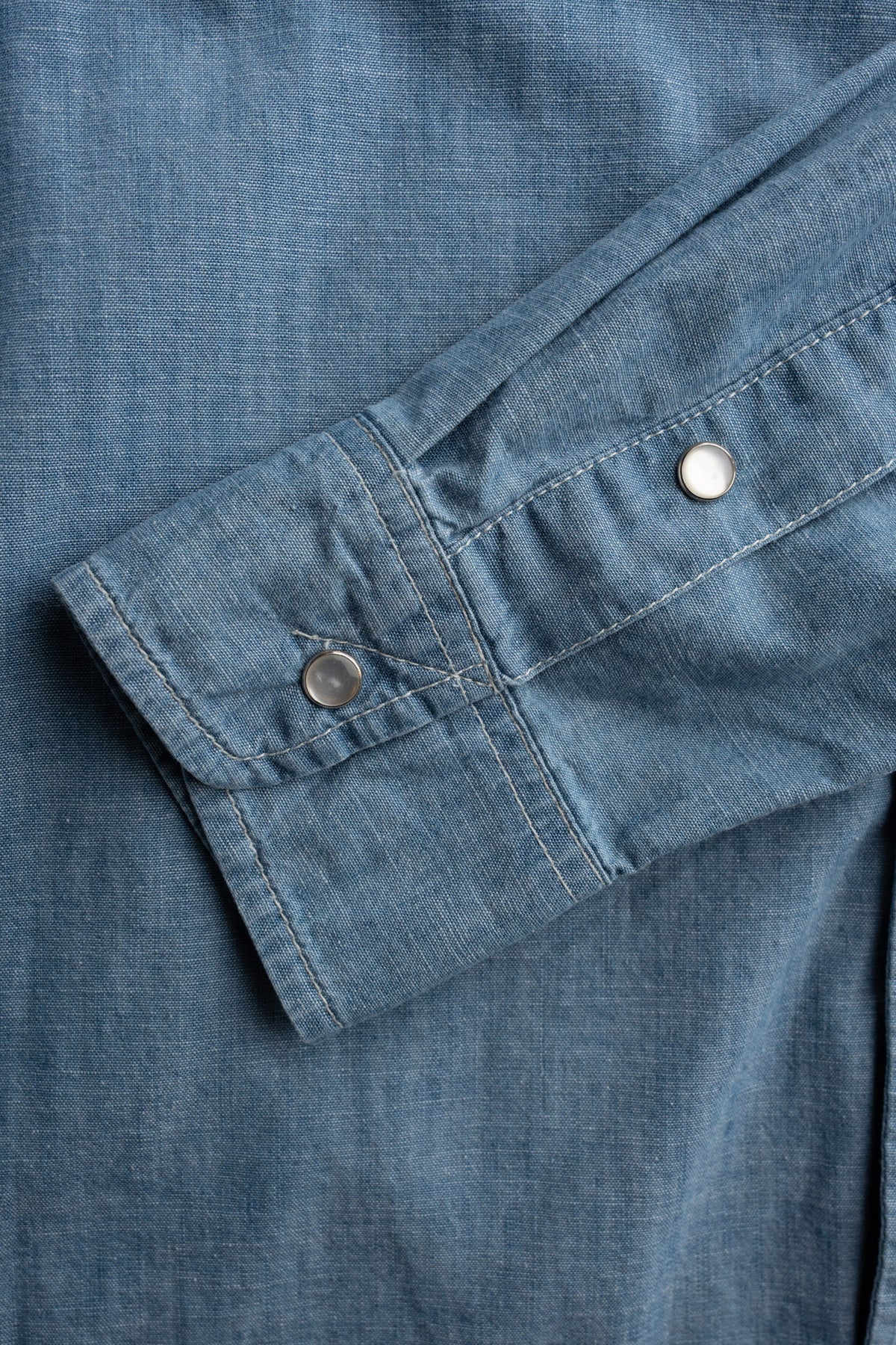 Close-up of a blue denim shirt sleeve with two metal snap buttons, highlighting the fabric texture and stitching details.