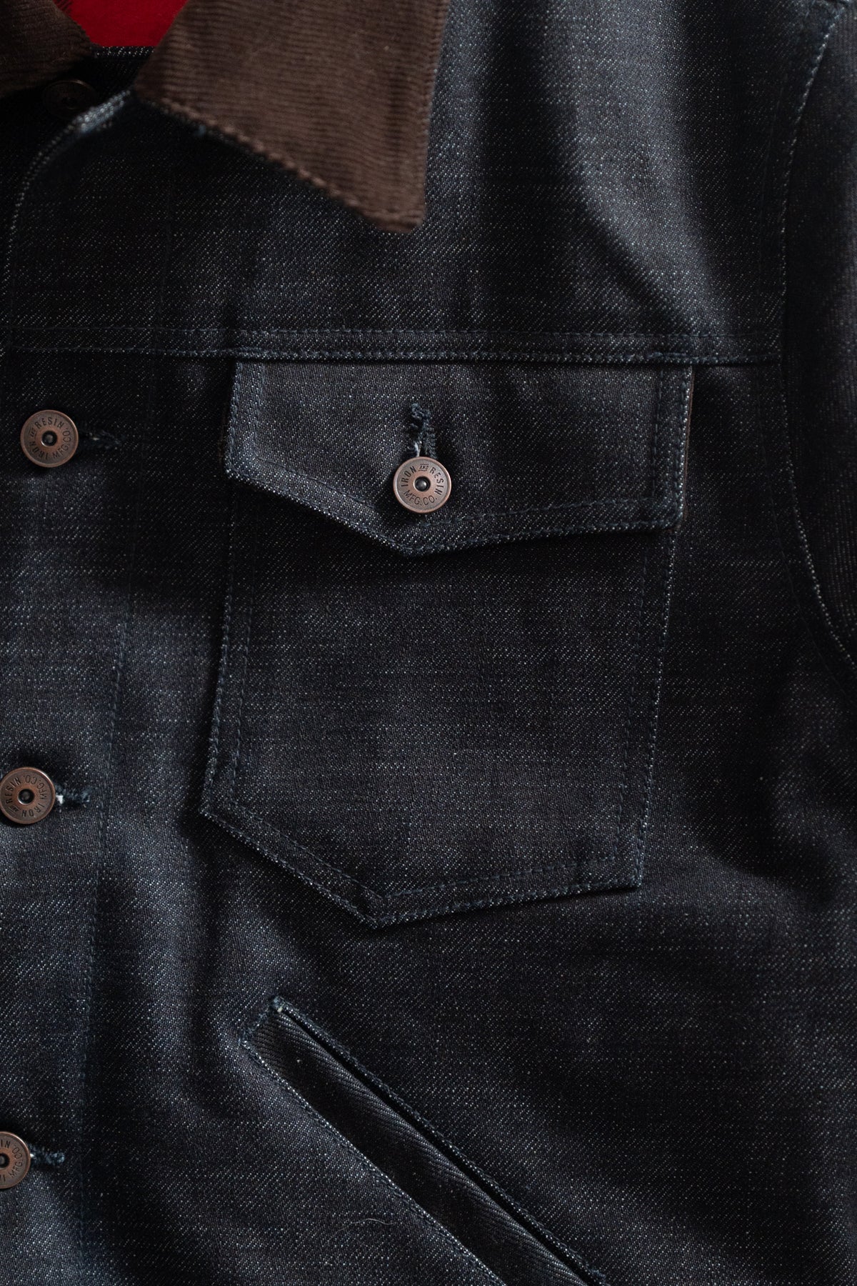 Close-up of a dark denim jacket with a brown collar, featuring metal buttons and a chest pocket, highlighting texture and design.