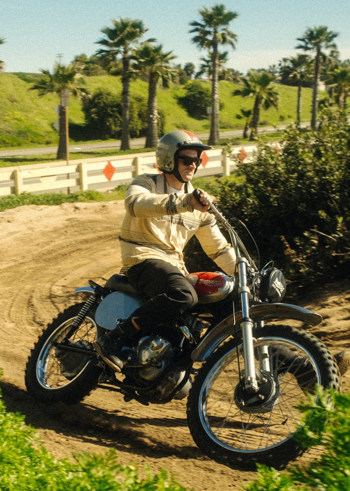 Man rides vintage motorcycle on a dirt path, surrounded by palm trees and greenery, under a clear blue sky.