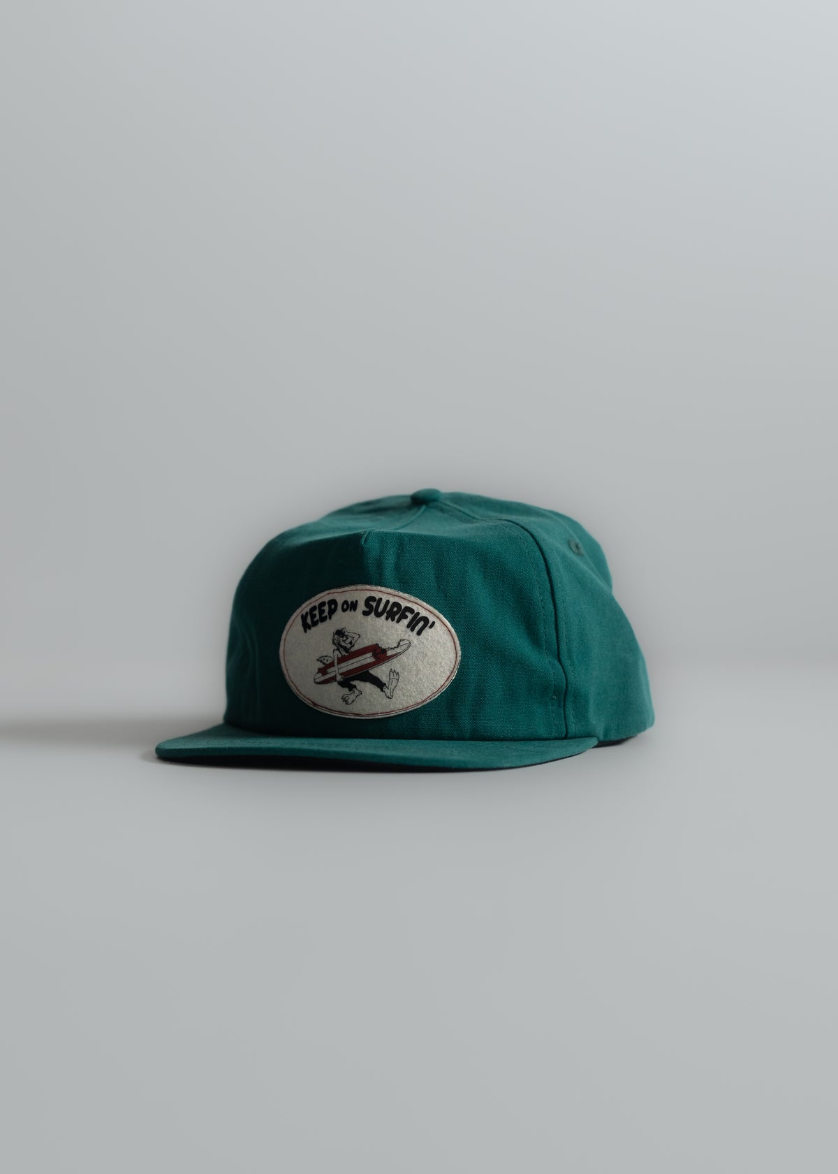 Green cap with "Keep on Surfin'" patch featuring a cartoon surfer, set against a plain gray background.