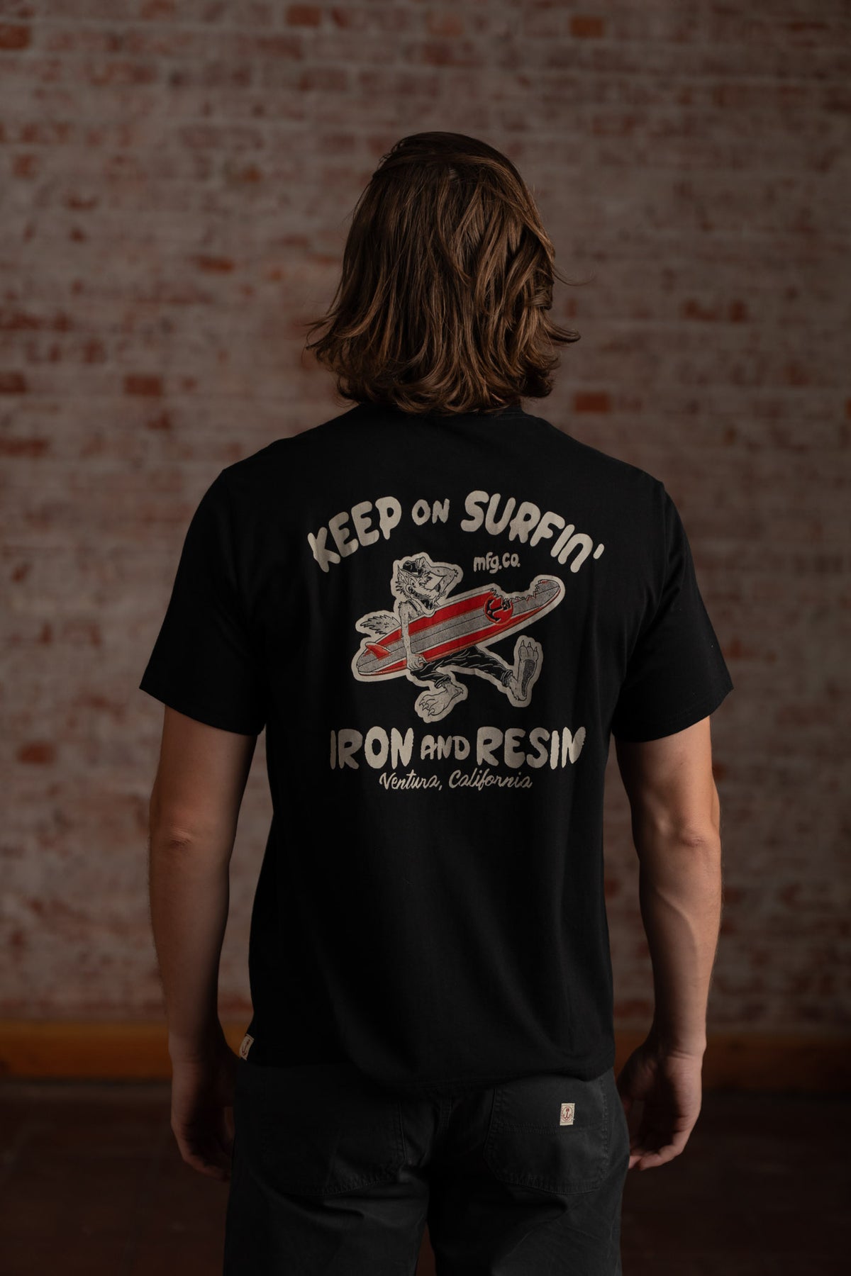 Man in black shirt with "Keep on Surfin'" and "Iron and Resin, Ventura, California" text, stands against a brick wall.