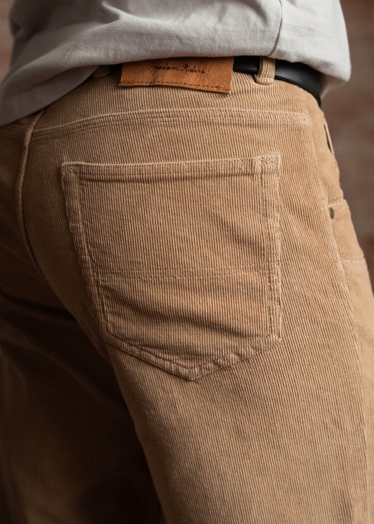 Close-up of tan corduroy pants with a visible back pocket and a "Freedom Riders" leather label, worn with a tucked white shirt.