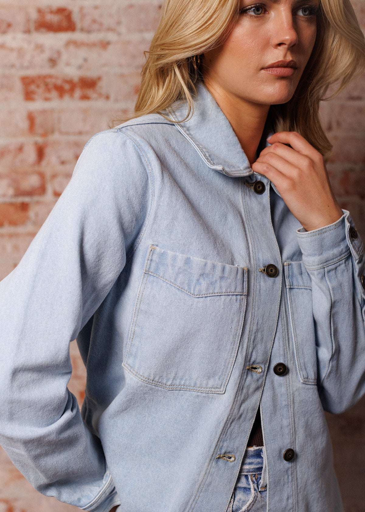 Woman in a light denim jacket stands against a brick wall, gazing thoughtfully. Her hand rests near her collar.