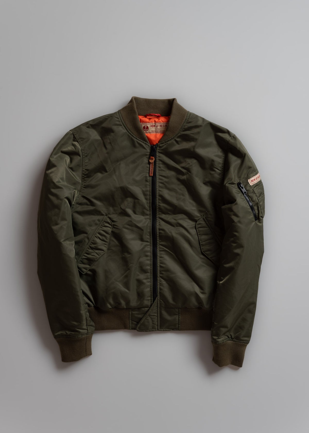 Olive green bomber jacket with orange lining, front zipper, and sleeve pocket, displayed on a gray background.