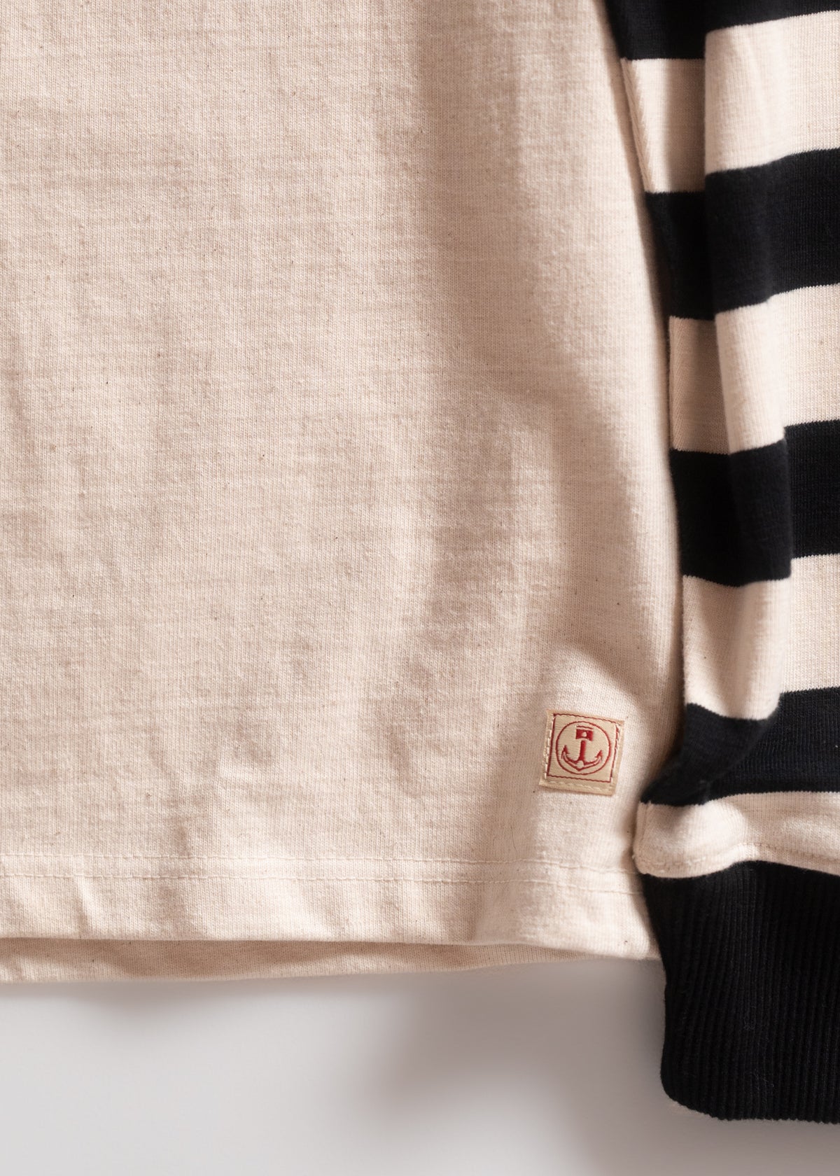 Beige shirt with black stripes and a red anchor patch on the hem. The fabric texture is visible, highlighting its quality.