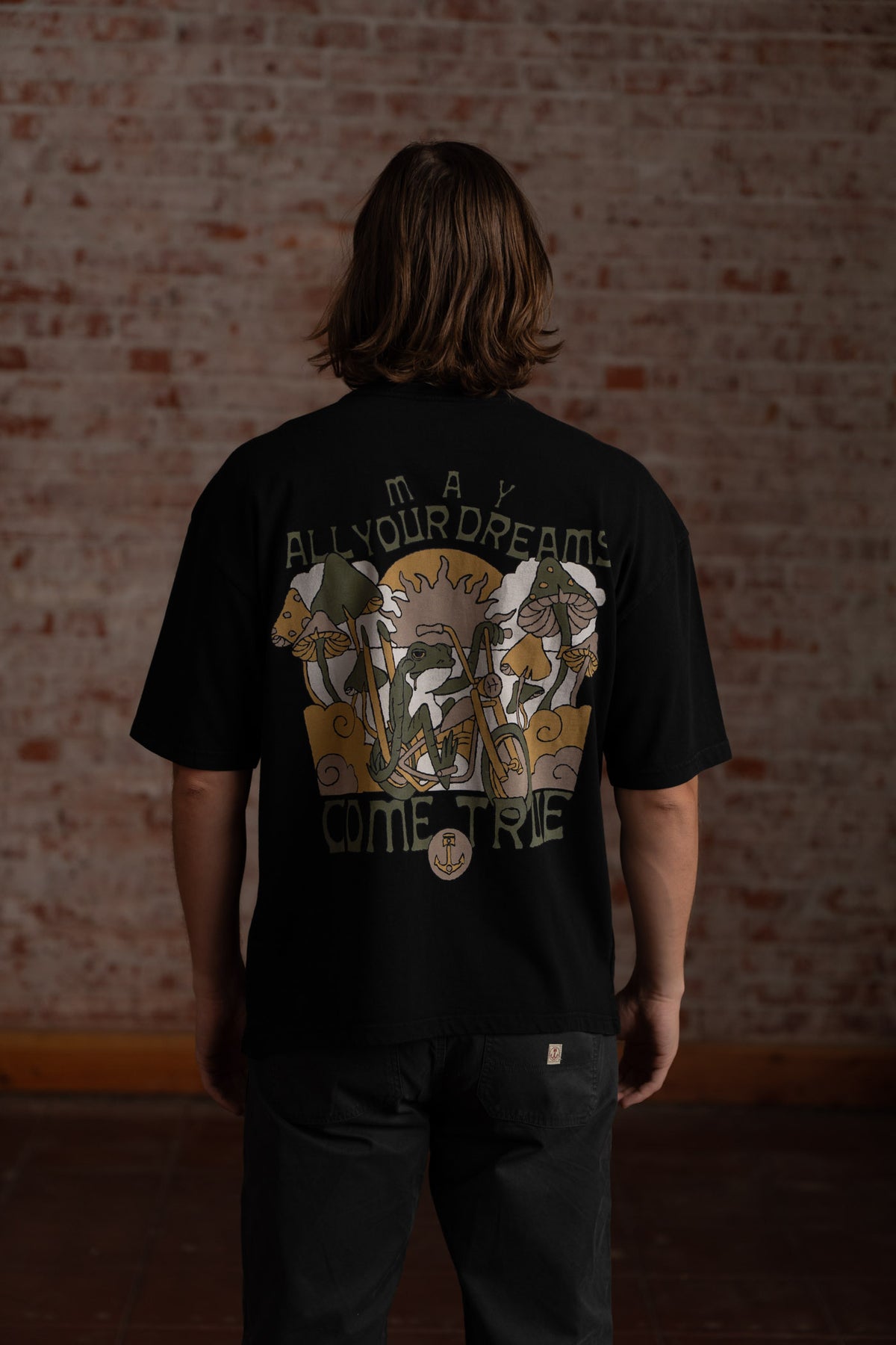 Man in black shirt with "May All Your Dreams Come True" text and mushroom design, standing against a brick wall background.
