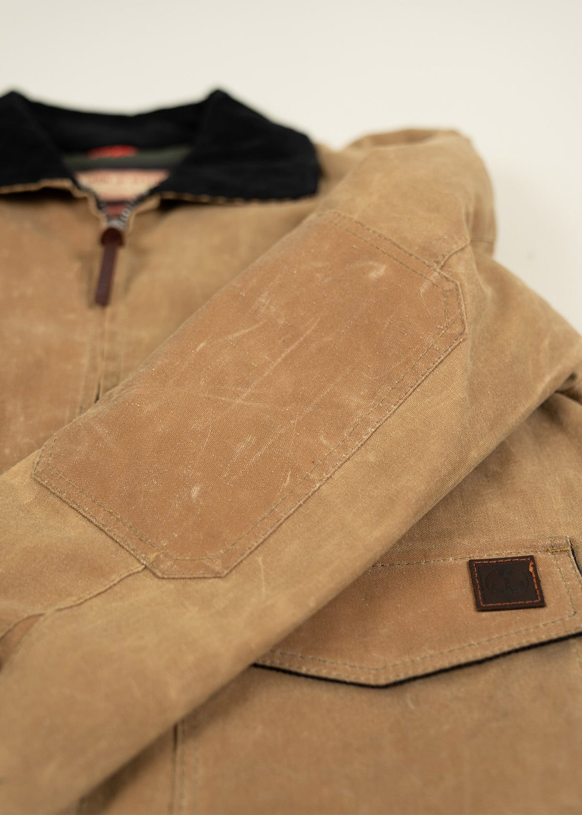 Close-up of a tan waxed canvas jacket with a black collar, highlighting the rugged texture and elbow patch detail.