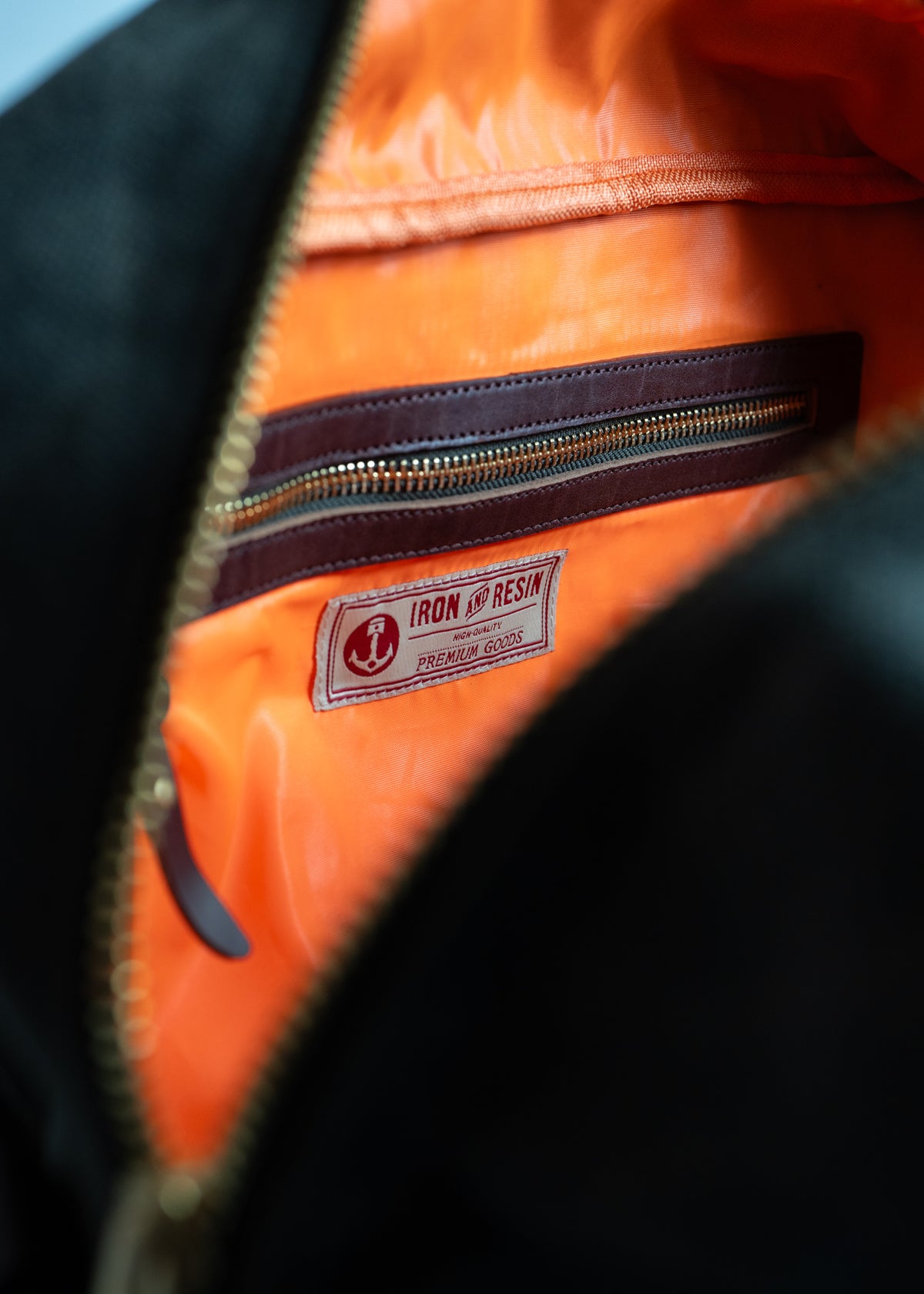 Open black bag reveals orange interior with a label reading "Iron and Resin Premium Goods" and a brown zippered pocket.