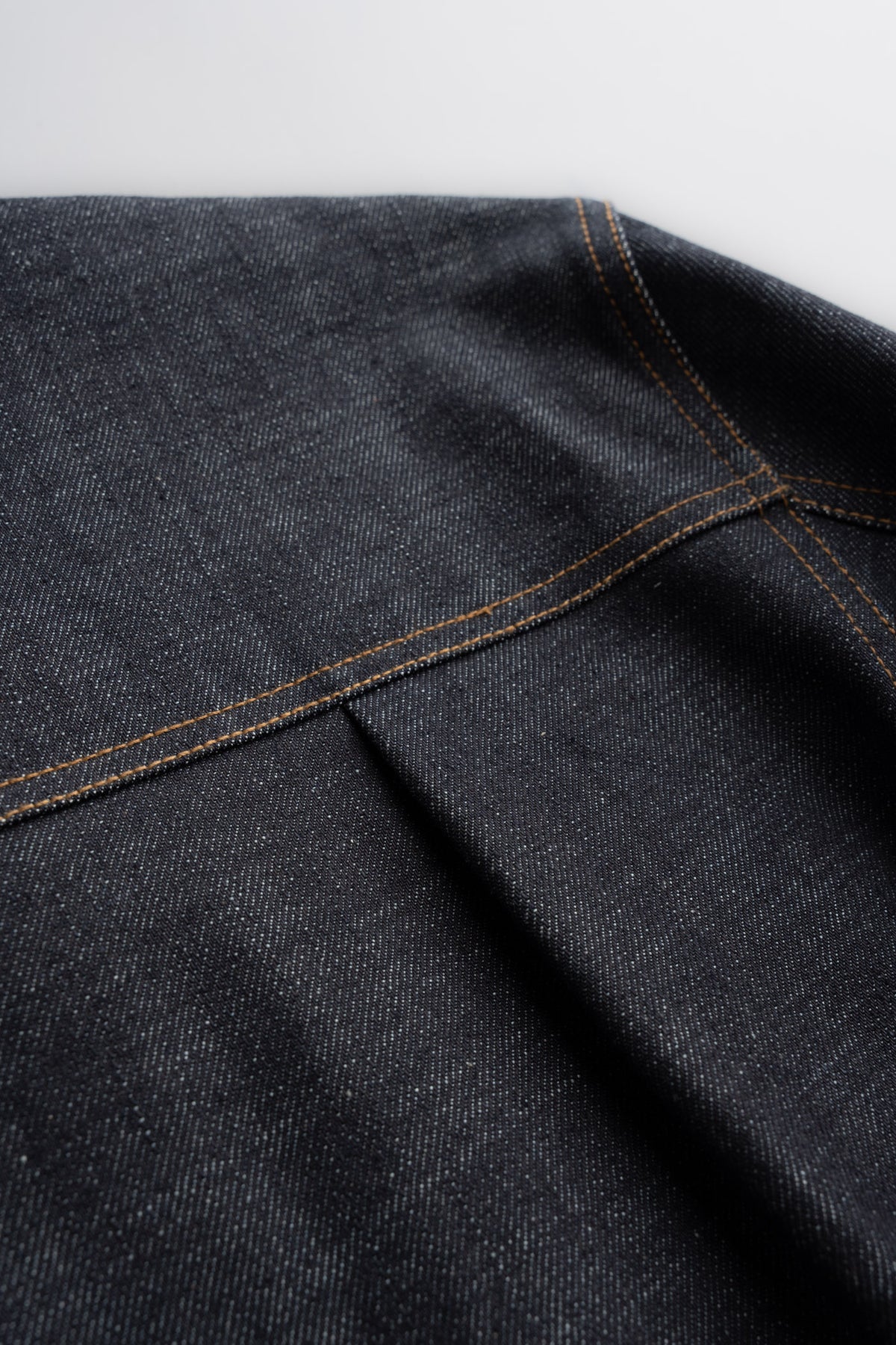 Close-up of dark denim fabric with visible orange stitching and a seam, showcasing texture and craftsmanship in detail.