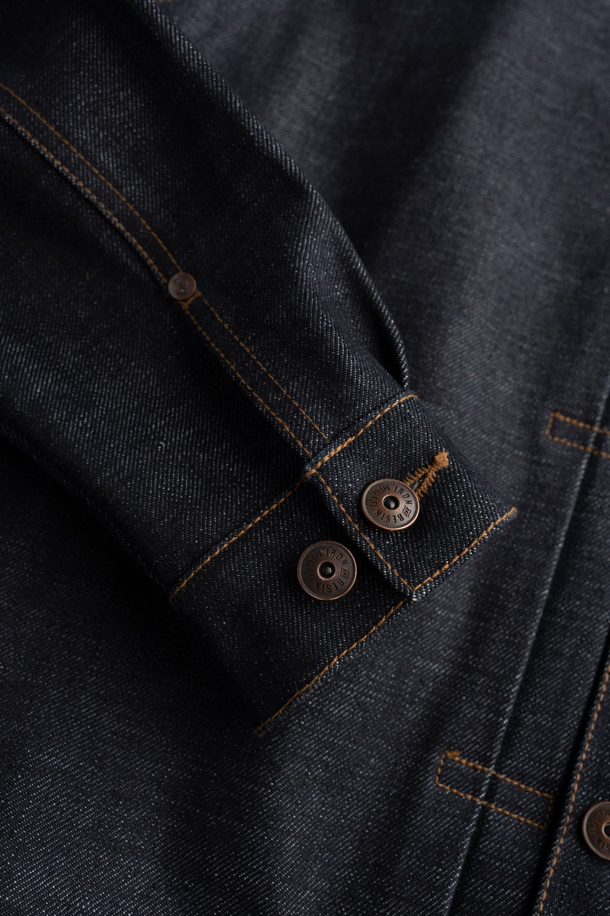 Close-up of a dark denim jacket sleeve with two metal buttons and orange stitching, highlighting the fabric texture and design.