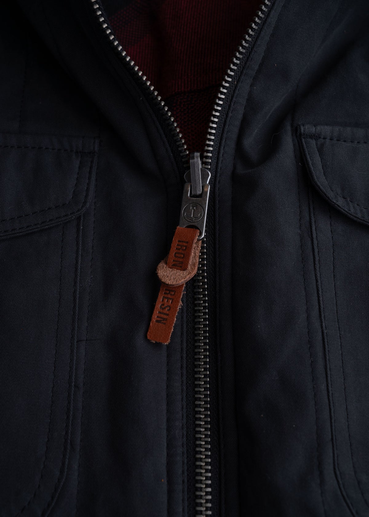 Close-up of a black jacket zipper with a brown leather pull tab labeled "Iron Resin," revealing a red plaid lining inside.
