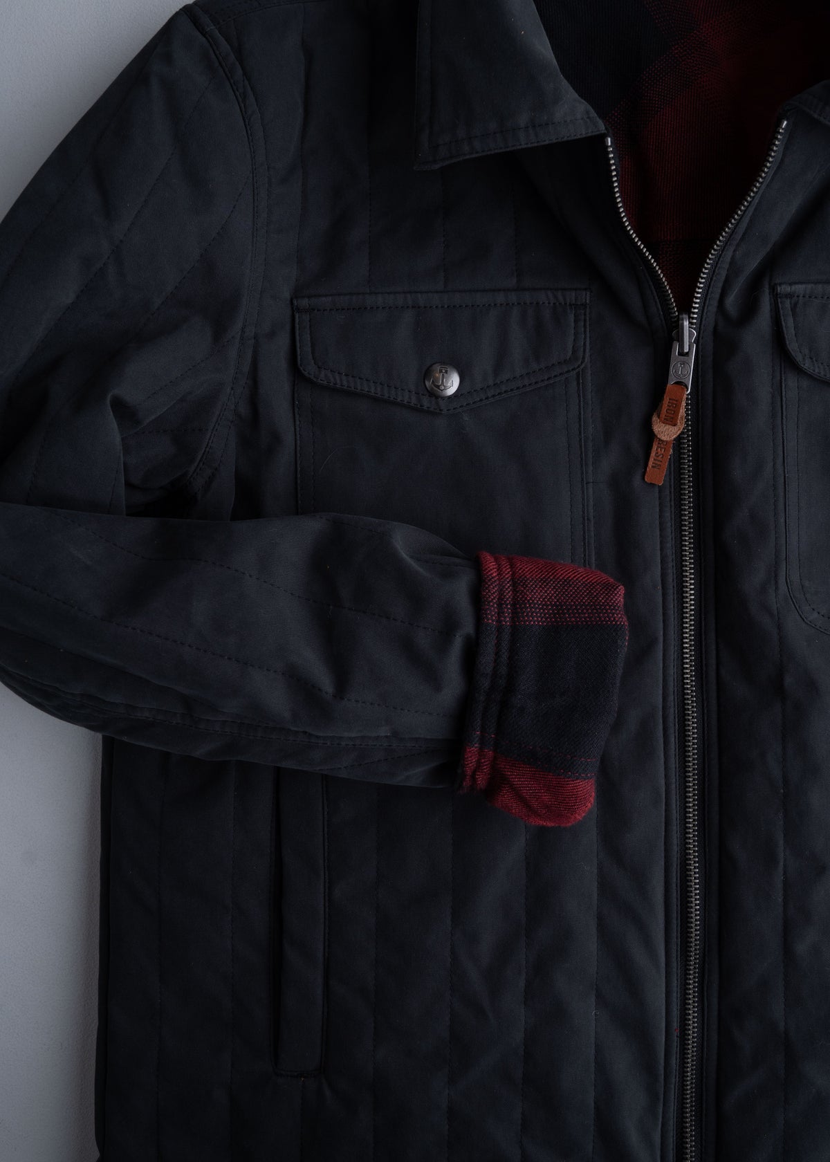 Black quilted jacket with a front zipper and buttoned pocket, featuring a red plaid lining peeking from the sleeve.