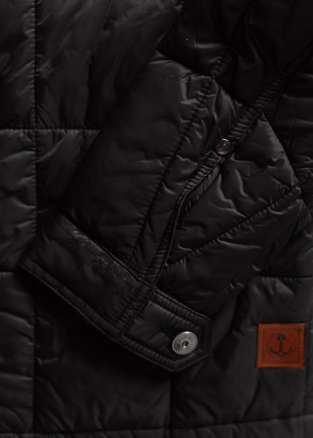 Close-up of a black quilted jacket with a visible button and brown anchor patch, highlighting its texture and design details.