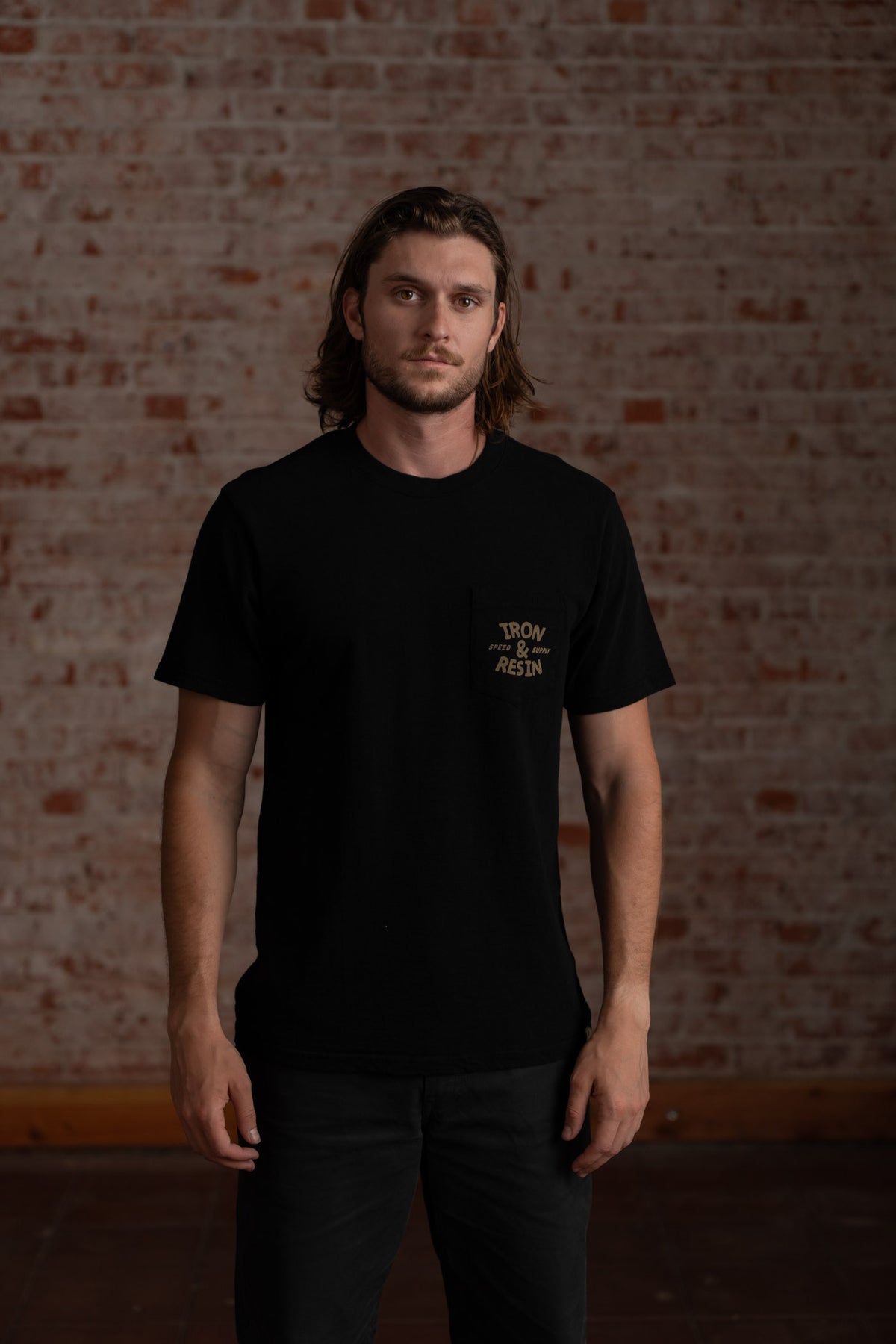 Man in black "Iron & Resin" shirt stands against a brick wall, exuding a casual, confident vibe.