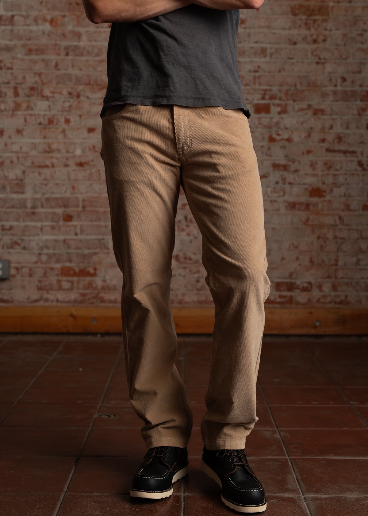 Man in beige pants and black shoes stands against a brick wall, arms crossed, wearing a dark shirt.