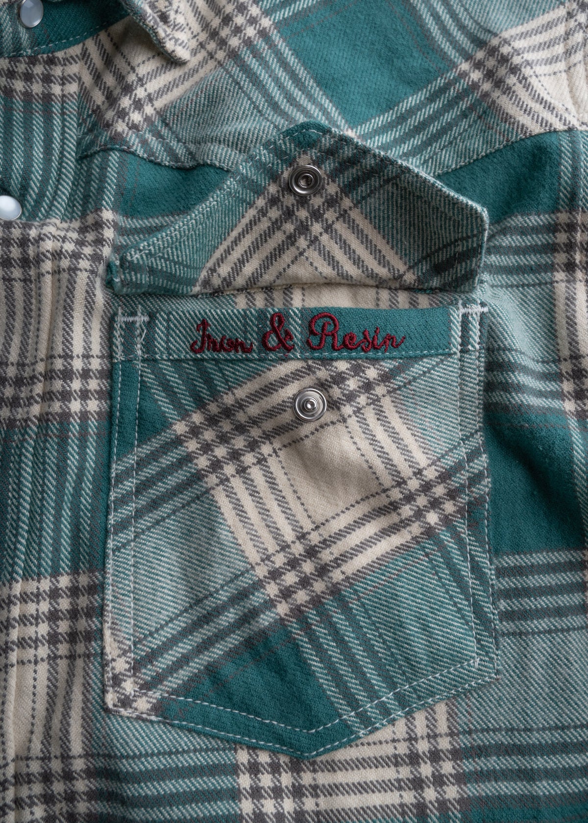 Close-up of a green plaid shirt pocket with "Iron & Resin" embroidered in red above a metal snap button.