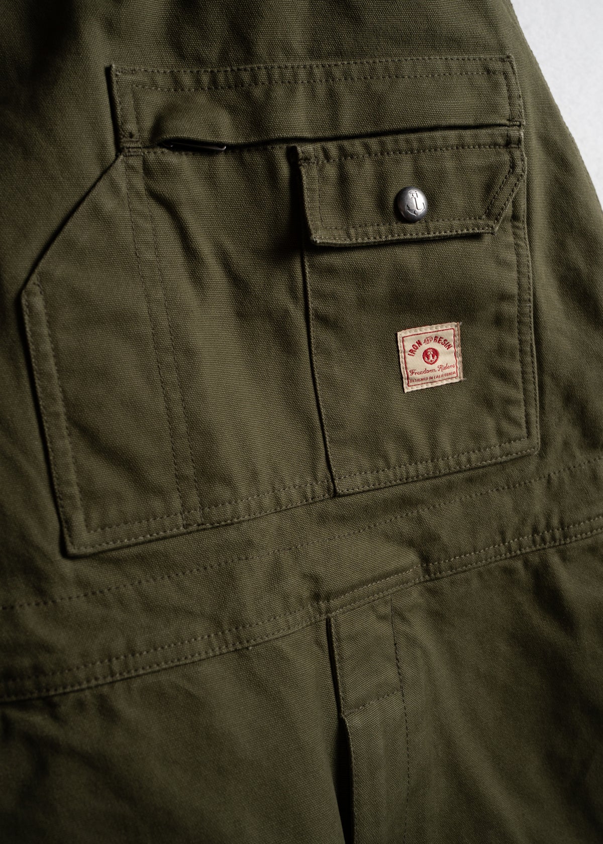 Olive green fabric with a buttoned flap pocket and a small red and white label featuring a logo. Durable and utilitarian design.