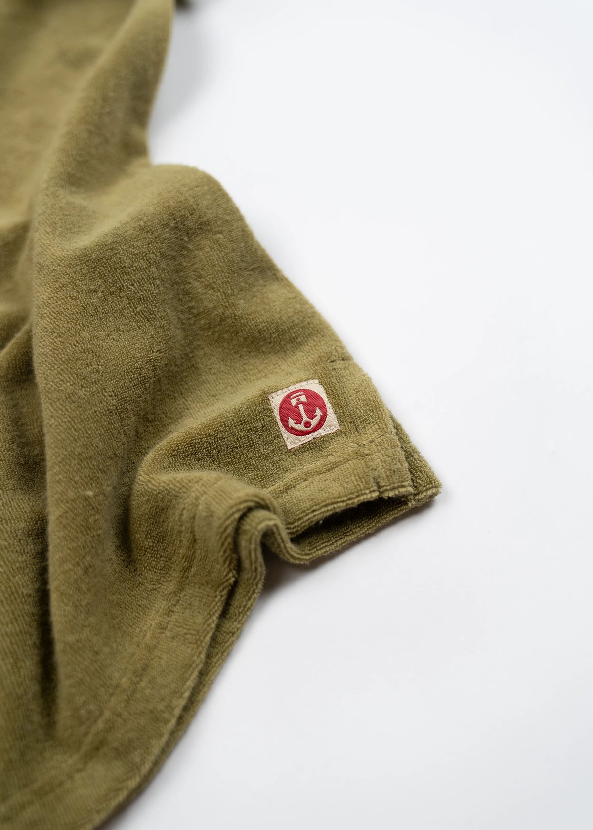 Olive green towel with a red anchor logo on a white tag, draped on a white surface, highlighting its soft texture.