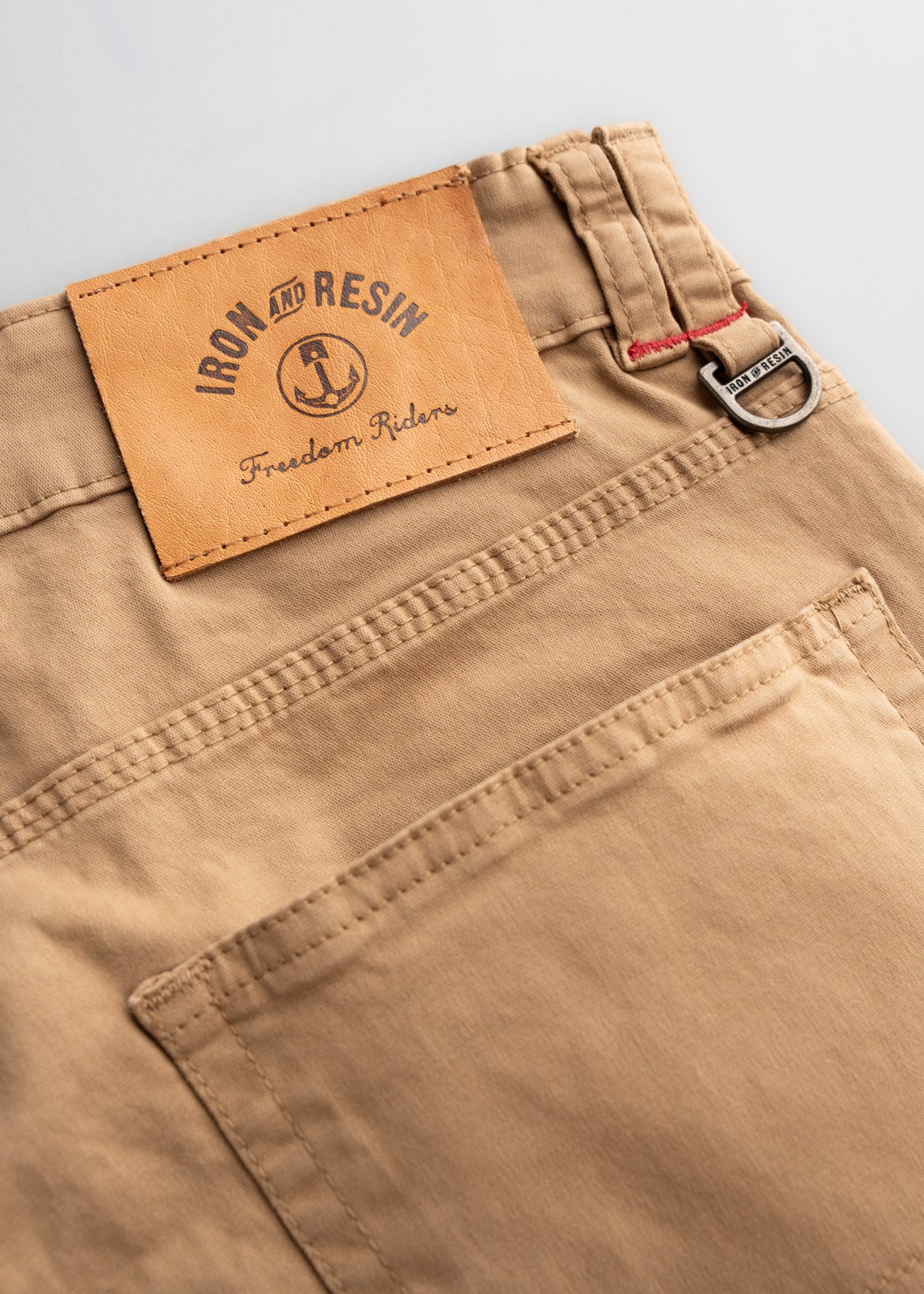 Close-up of tan pants with "Iron and Resin" leather patch and metal buckle detail, showcasing craftsmanship and style.