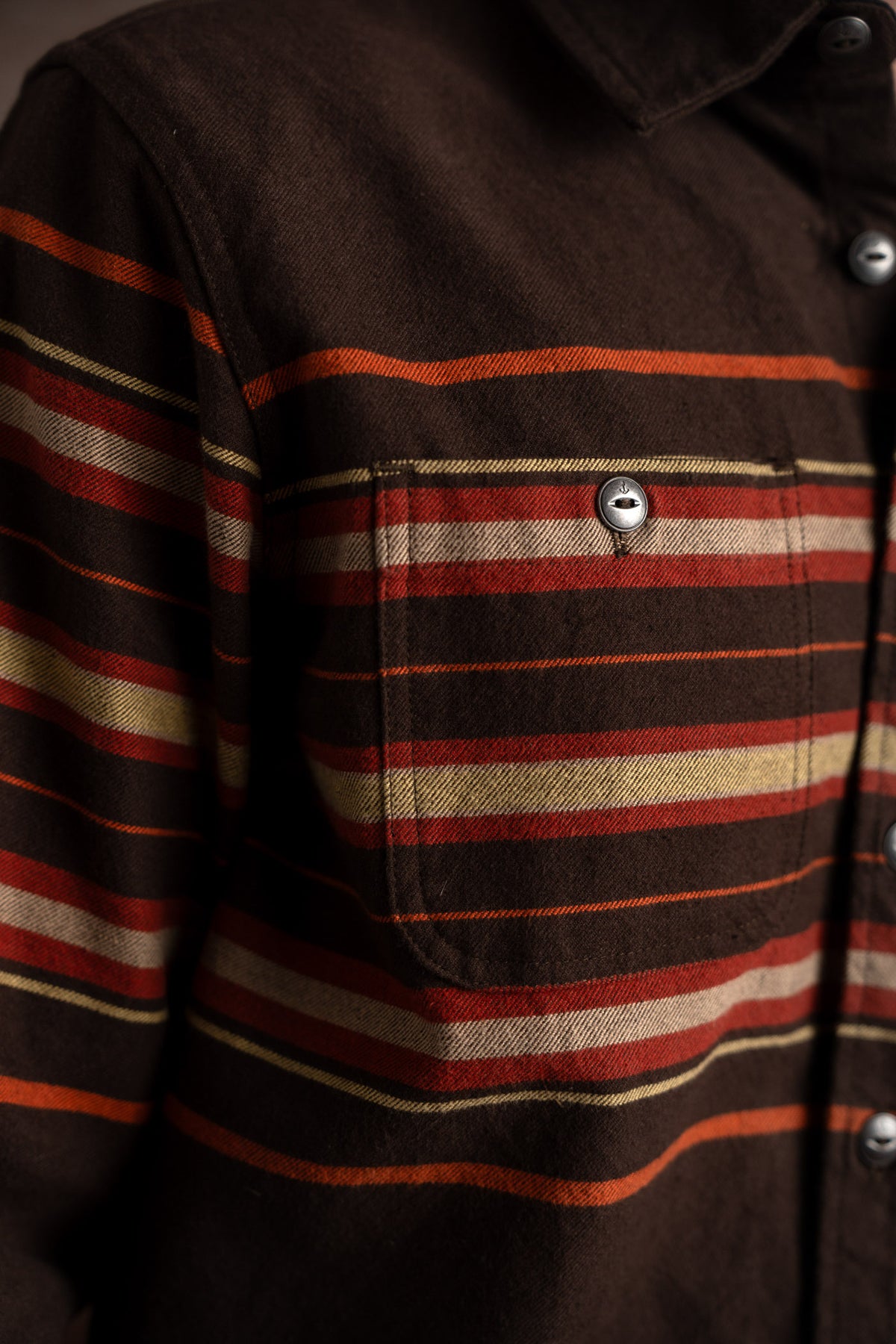 Close-up of a brown shirt with red, orange, and cream stripes, featuring a buttoned chest pocket and metal buttons.
