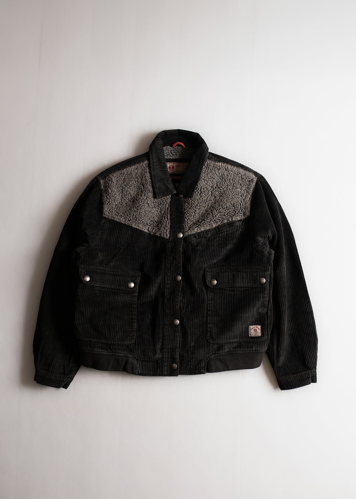 Black corduroy jacket with gray fleece yoke, buttoned pockets, and collar, displayed on a white background.