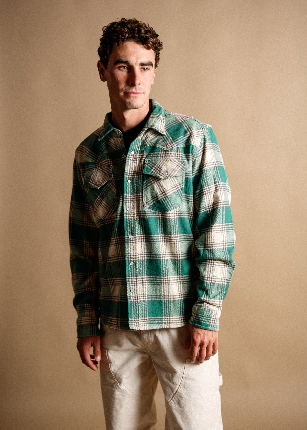 Man in green plaid shirt and white pants stands against a beige background, looking to the side with a thoughtful expression.