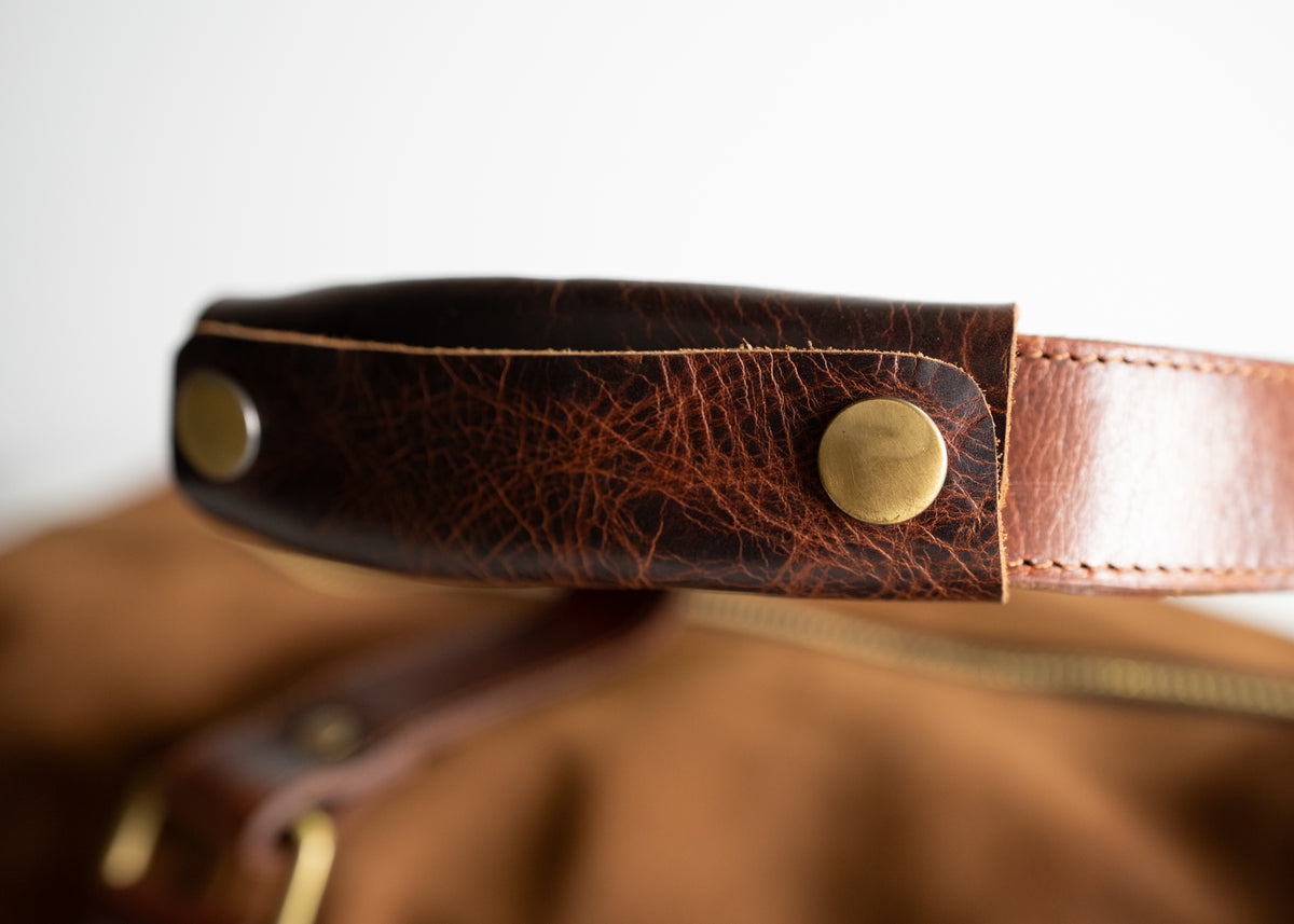 Close-up of a brown leather bag handle with brass rivets, showcasing detailed texture and craftsmanship against a soft background.