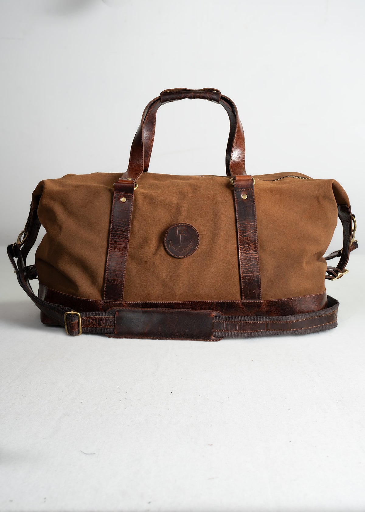 Brown canvas duffel bag with leather handles and strap on a white background, featuring a round logo patch in the center.